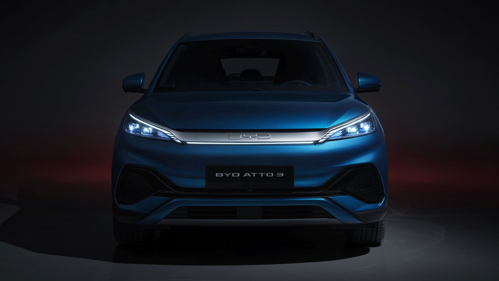 Frontansicht des BYD ATTO 3 in blauer Lackierung, durchgezogene LED-Scheinwerfer leuchten vor dem dunklen Hintergrund.