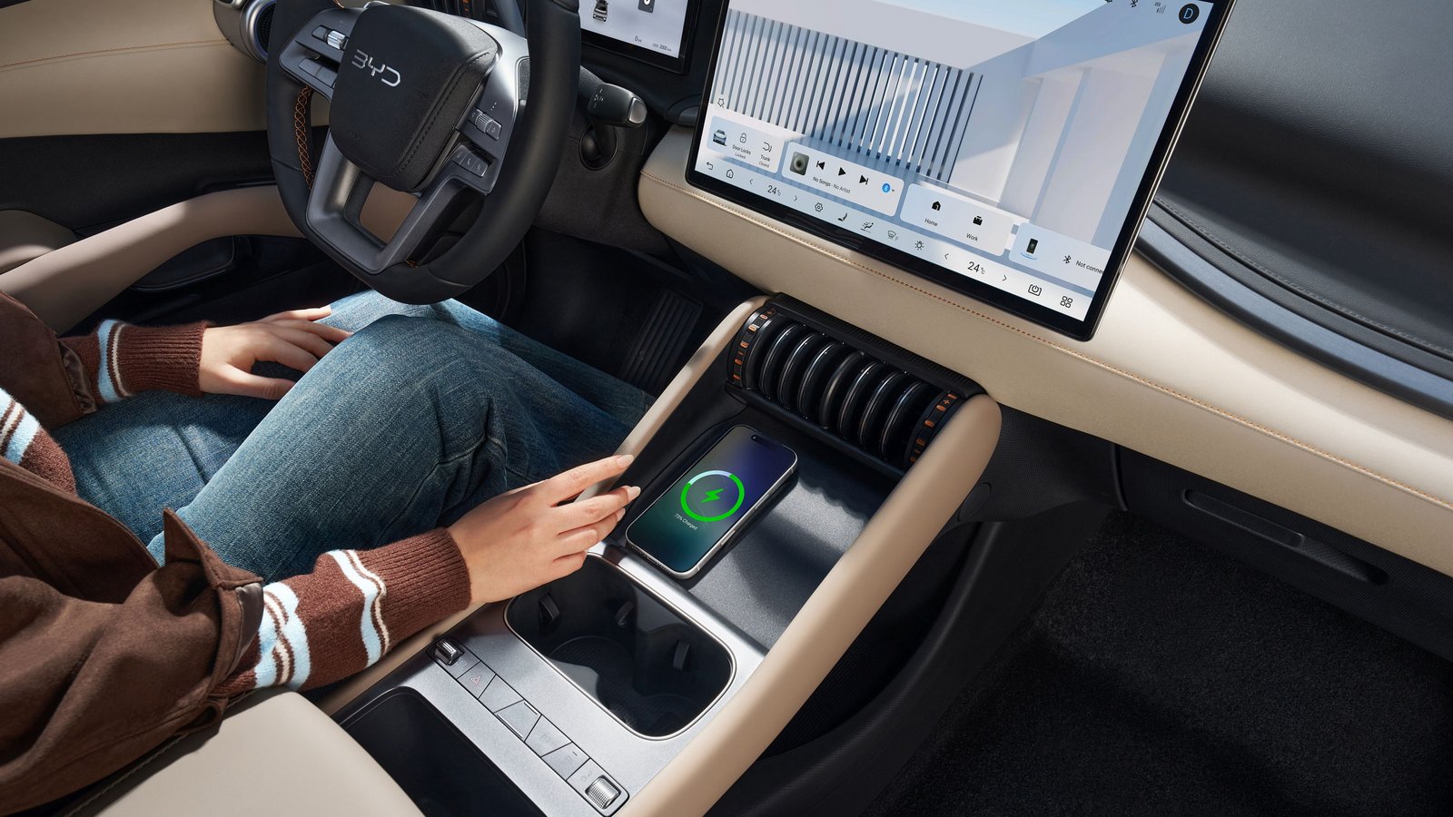 Innenraum des BYD ATTO 3 EVO mit beige-schwarzem Armaturenbrett, großem Touchscreen, kabelloser Smartphone-Ladestation und Person mit braunem Pullover und Jeans auf dem Fahrersitz