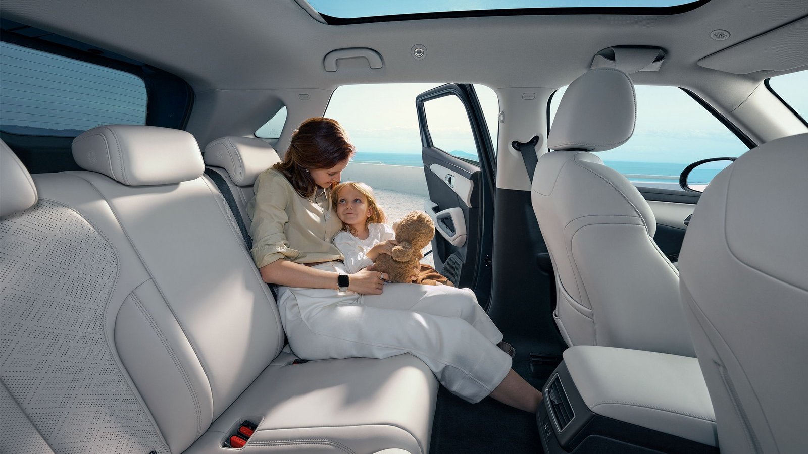 Innenraum eines BYD ATTO 2 DM-i mit hellen Sitzen, Panoramadach und geöffneter Tür, eine Person sitzt auf der Rückbank mit einem Kind und einem Teddybär