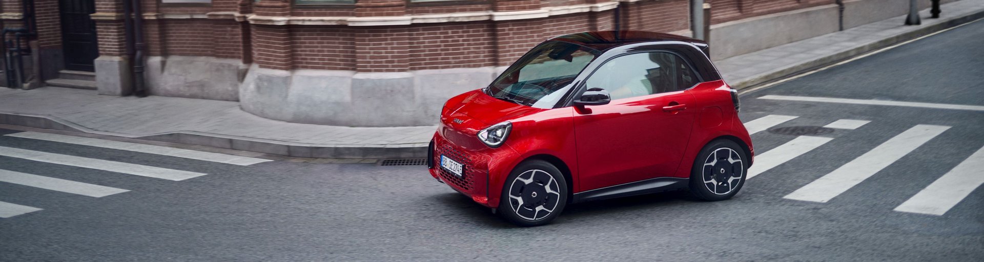 Roter smart #2 Kleinwagen mit schwarzem Dach fährt auf einer Kreuzung vor einem Backsteingebäude