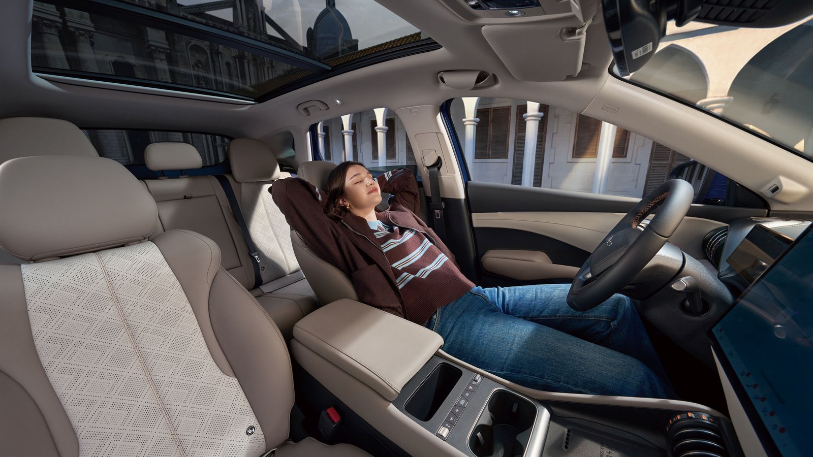 Innenraum des BYD ATTO 3 EVO mit Panoramadach, einer Person, die auf dem Beifahrersitz entspannt zurücklehnt, und sichtbarem Armaturenbrett mit großem Touchscreen.