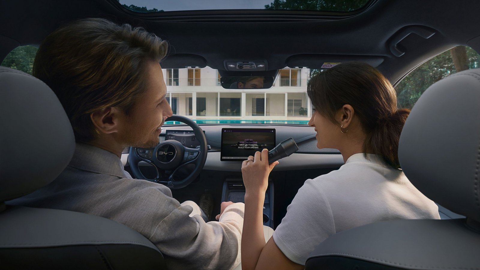 Innenansicht eines Autos mit zwei Personen auf den Vordersitzen. Eine Person hält ein Mikrofon, während der Bildschirm in der Mittelkonsole Informationen anzeigt.